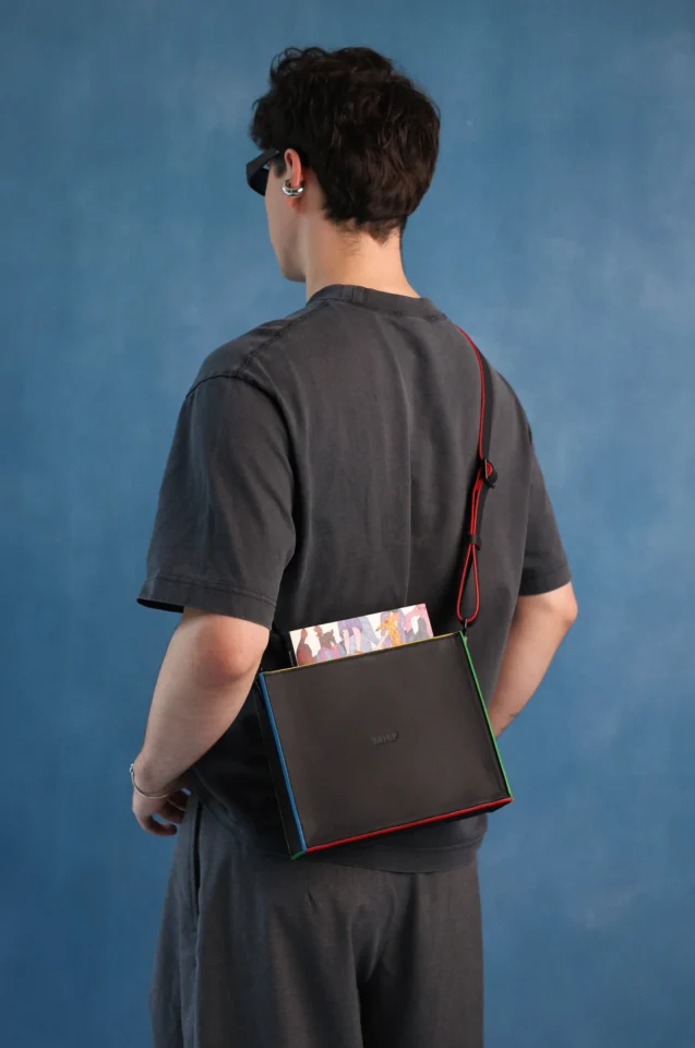 A person shown from the back wearing a dark gray t-shirt and shorts, carrying a black crossbody bag with a red strap and colorful edges over their shoulder, set against a textured blue studio backdrop.