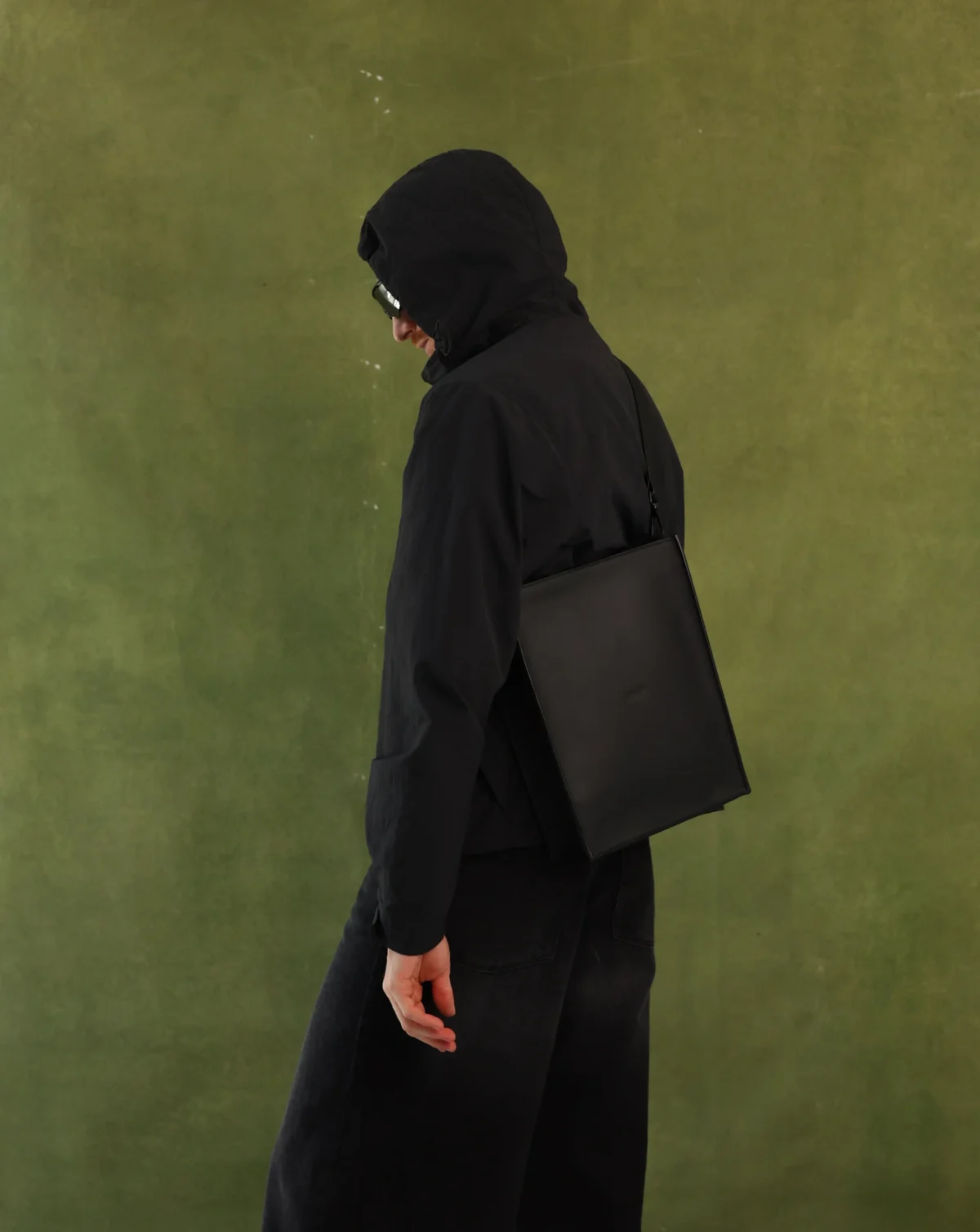 A person in a black hooded jacket and black pants, shown from the back and side, carrying a black leather shoulder bag against a textured olive green background.